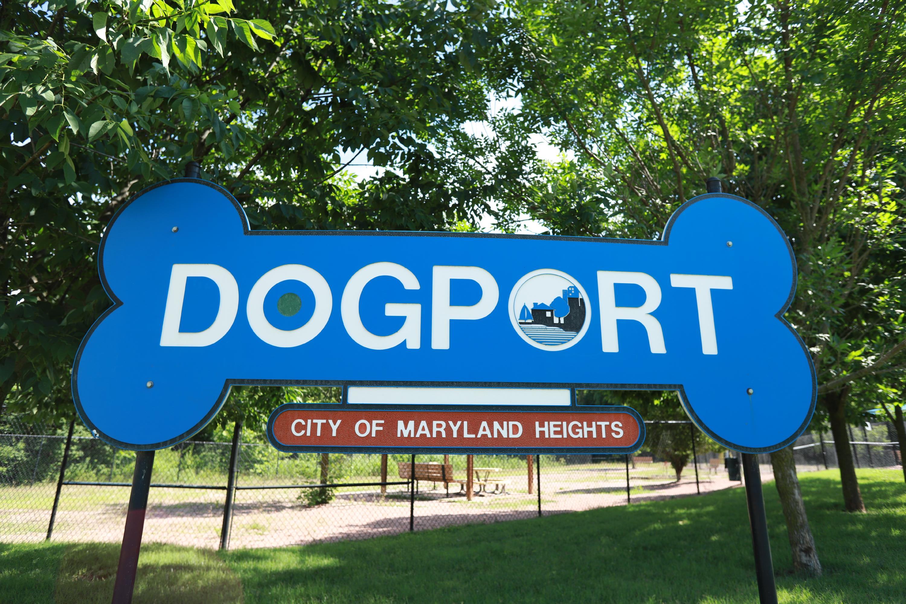 A blue sign in the shape of a dog bone contains the logo for the "Dogport" park, with the City of Maryland Heights logo replacing the second "O" of the park. In the background, green trees are in front of the sandy play area on a sunny summer day.